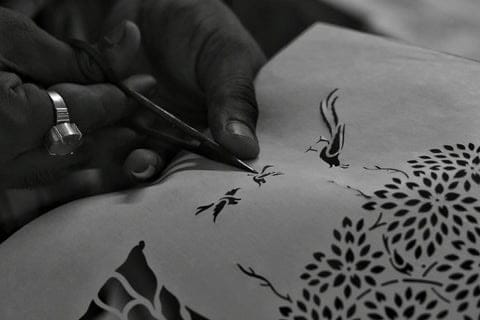 A Sanjhi artist cutting a paper stencil with fine curved scissors